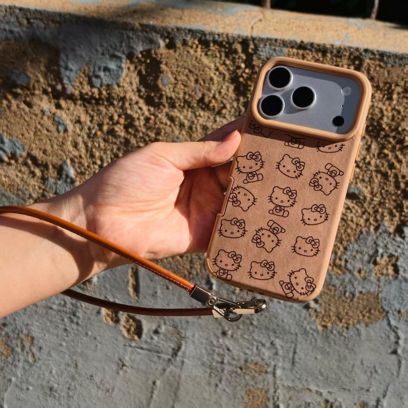 May include: A tan smartphone case featuring a repeating black line art pattern of the Hello Kitty character. The case includes a raised camera frame and a thin, brown leather-like wrist strap attached with a silver-toned metal clasp.