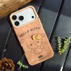 May include: A brown phone case featuring a Hello Kitty design. The case has a tan border around the camera and a yellow bow with a gem. The words "HELLO KITTY" are printed at the top, and the phrase "Hello Kitty can be happy, confident and happy" is at the bottom.