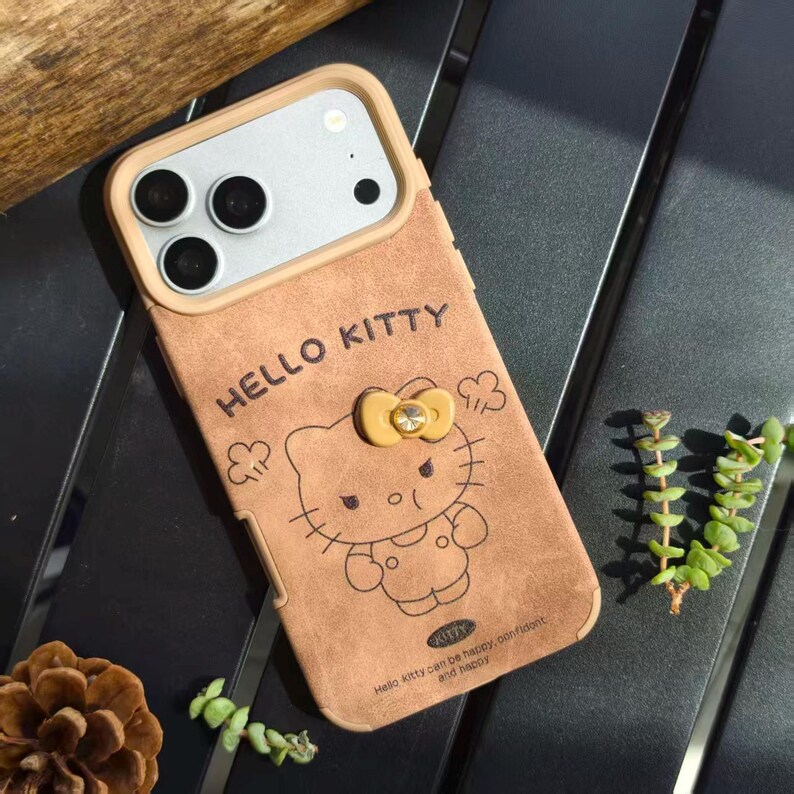 May include: A brown phone case featuring a Hello Kitty design. The case has a tan border around the camera and a yellow bow with a gem. The words "HELLO KITTY" are printed at the top, and the phrase "Hello Kitty can be happy, confident and happy" is at the bottom.
