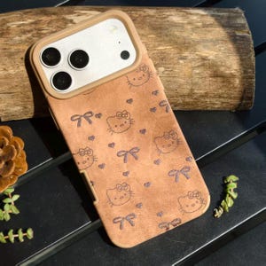 May include: A brown phone case featuring a repeating pattern of Hello Kitty faces, bows, and hearts. The case has a tan border around the camera and a matching tan frame. The case is on a dark surface.