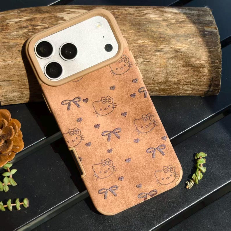 May include: A brown phone case featuring a repeating pattern of Hello Kitty faces, bows, and hearts. The case has a tan border around the camera and a matching tan frame. The case is on a dark surface.