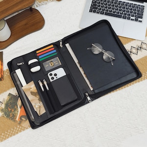 May include: A black zippered portfolio organizer featuring multiple interior slots for cards, a smartphone, two pens, and a white wireless earbud case. The right side contains a large document sleeve with a pair of round metal-framed glasses resting on top.
