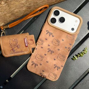 K&ouml;nnte beinhalten: Eine braune Handyh&uuml;lle und eine passende kleine Tasche mit einem sich wiederholenden Muster aus Hello Kitty-Gesichtern und Schleifen. Die Handyh&uuml;lle hat einen Kameraausschnitt und eine Handschlaufe. Die Tasche hat einen kleinen Kartenhalter.