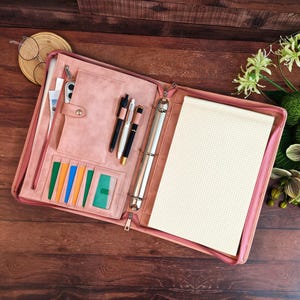May include: A dusty rose zippered portfolio organizer featuring a three-ring metal binder, a grid-lined notepad, multiple card slots, a vertical pocket, and three pen loops holding black and white pens. The interior is lined with matching fabric.