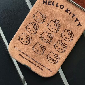 Op de afbeelding: Bruine telefoonhoes met meerdere Hello Kitty-illustraties. De woorden "HELLO KITTY" staan bovenaan en een citaat onderaan. De hoes is gemaakt van een textuurrijk materiaal.