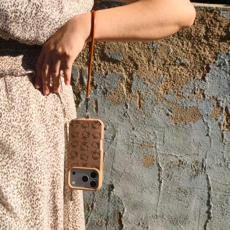 May include: A tan smartphone case featuring a repeating pattern of Hello Kitty character faces in black outlines. The case includes a matching tan wrist strap attached to a silver metal loop at the top, designed for carrying the mobile device.