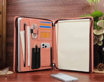 May include: A salmon-colored zippered portfolio organizer featuring a laptop sleeve, a vertical zippered pocket, multiple card slots, a clear ID window, two pen loops, and a large grid-lined notepad. A smartphone rests on the surface in front.