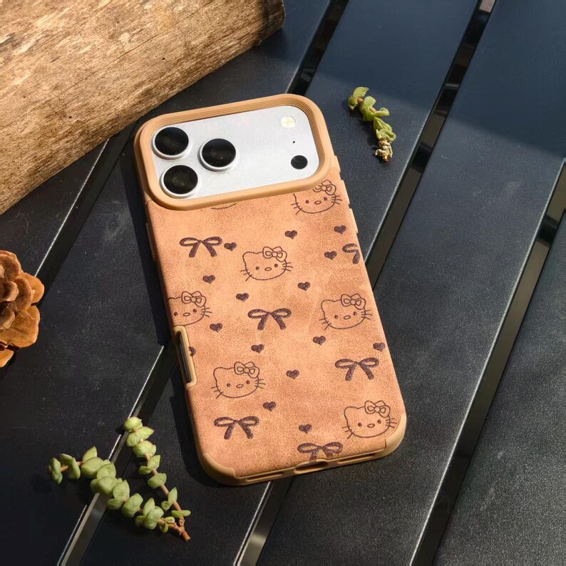 May include: A tan phone case featuring a repeating pattern of Hello Kitty faces, bows, and hearts. The case has a brown border and a camera cutout. The case is made of a textured material.