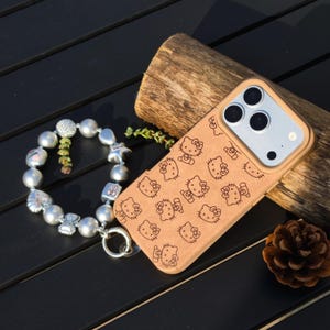 May include: A tan phone case featuring a repeating pattern of Hello Kitty faces. A silver charm bracelet with various charms and large pearl beads is next to the phone case. A small pine cone and a piece of wood are also in the image.
