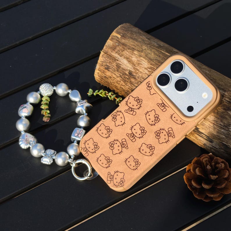 May include: A tan phone case featuring a repeating pattern of Hello Kitty faces. A silver charm bracelet with various charms and large pearl beads is next to the phone case. A small pine cone and a piece of wood are also in the image.