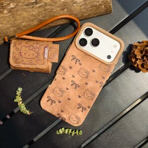 May include: A brown phone case and small pouch featuring a repeating Hello Kitty pattern with bows and hearts. The phone case has a tan border around the camera. A brown wrist strap is attached to the pouch.