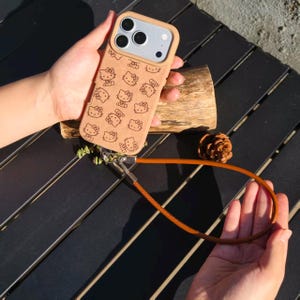 May include: A tan phone case featuring a repeating pattern of black Hello Kitty character outlines. The case includes a camera cutout and a long, thin brown leather wrist strap attached to the bottom with a silver metal clasp and hardware.