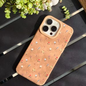 May include: A tan phone case with a floral pattern featuring small red, blue, and white flowers. The case has a beige camera ring and a soft, textured appearance. The case is designed for a smartphone.