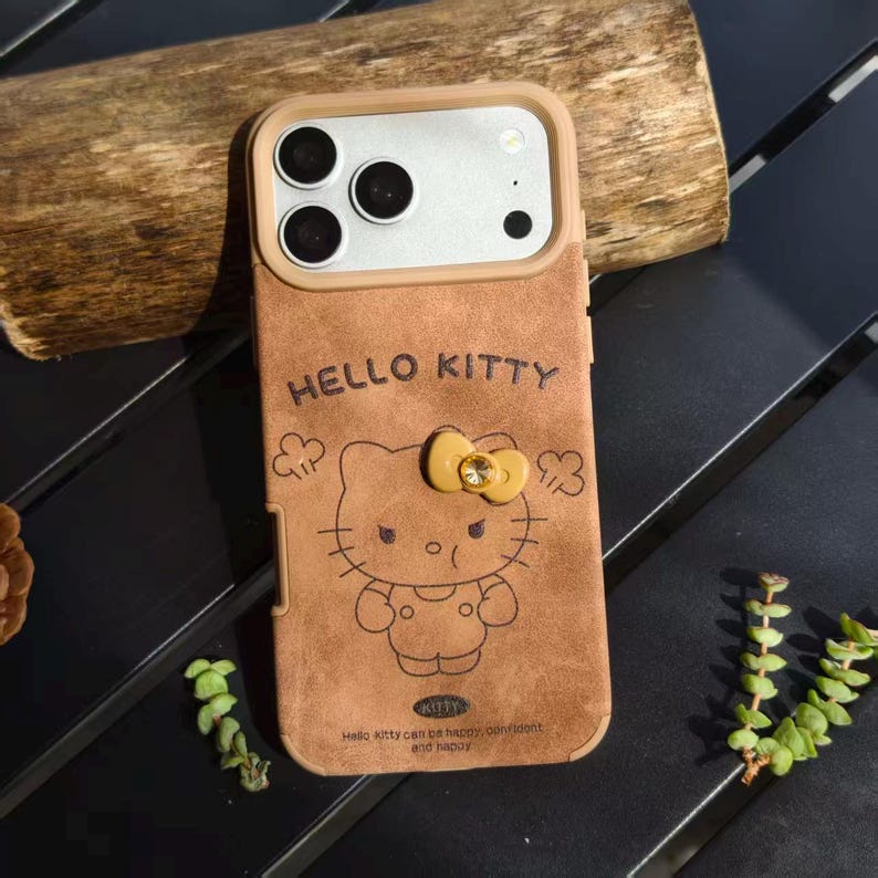 May include: Brown phone case featuring a Hello Kitty design. The case has a tan border around the camera and a bow with a jewel. The text "HELLO KITTY" is above the character, and the text "Hello kitty can be happy, confident and happy" is below.