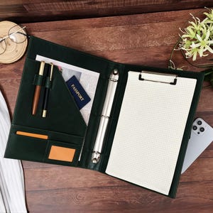 May include: A dark green leather-like portfolio binder featuring a metal three-ring mechanism, a clipboard clip, two pen loops, and interior pockets. The left side holds two pens, a document, and a blue passport. The right side contains a grid paper pad.
