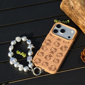 May include: A tan phone case featuring a repeating pattern of Hello Kitty characters. A silver charm bracelet with various charms and beads is next to the phone case. The bracelet includes a star charm with the word "PINK".