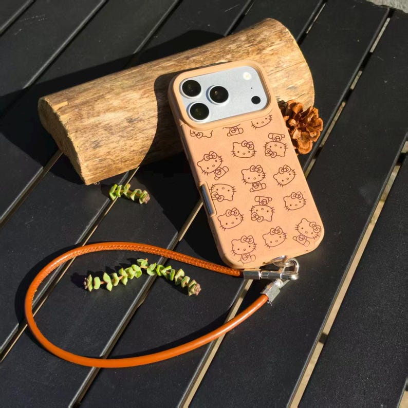 May include: A tan smartphone case featuring a repeating black line art pattern of the Hello Kitty character. The case includes a silver-toned camera lens frame and a detachable orange leather wrist strap attached with silver metal hardware.