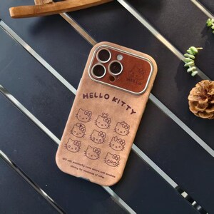 Op de afbeelding: Een bruine telefoonhoes met de tekst "HELLO KITTY" en meerdere illustraties van Hello Kitty's gezicht. De hoes heeft een bruine leerachtige textuur en een bruin camerakader. De onderkant van de hoes heeft een citaat.