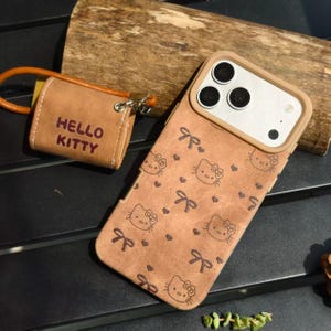 May include: A brown phone case with a Hello Kitty pattern, featuring bows and hearts. A matching brown accessory with "HELLO KITTY" printed on it is attached with a brown strap. The phone case has a camera cutout.