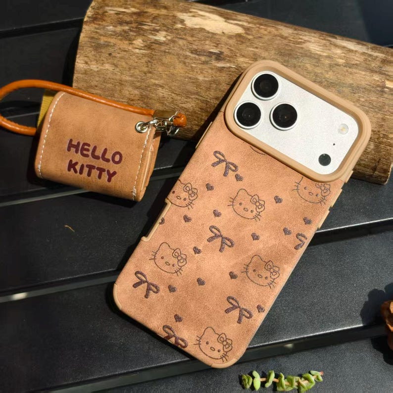 May include: A brown phone case with a Hello Kitty pattern, featuring bows and hearts. A matching brown accessory with "HELLO KITTY" printed on it is attached with a brown strap. The phone case has a camera cutout.