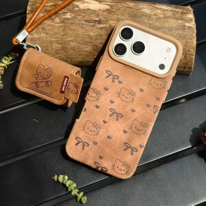K&ouml;nnte beinhalten: Braune Handyh&uuml;lle und AirPod-H&uuml;lle mit Hello Kitty-Muster. Die Handyh&uuml;lle hat einen braunen Rahmen um die Kameralinsen. Die AirPod-H&uuml;lle hat einen braunen Lederriemen. Die H&uuml;llen liegen auf einer Holzoberfl&auml;che.