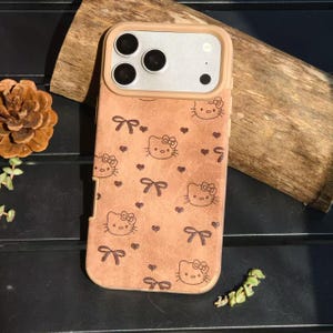 May include: A tan phone case featuring a Hello Kitty design with bows and hearts. The case has a tan frame around the camera and a light brown back. The Hello Kitty faces and bows are in a dark brown color.