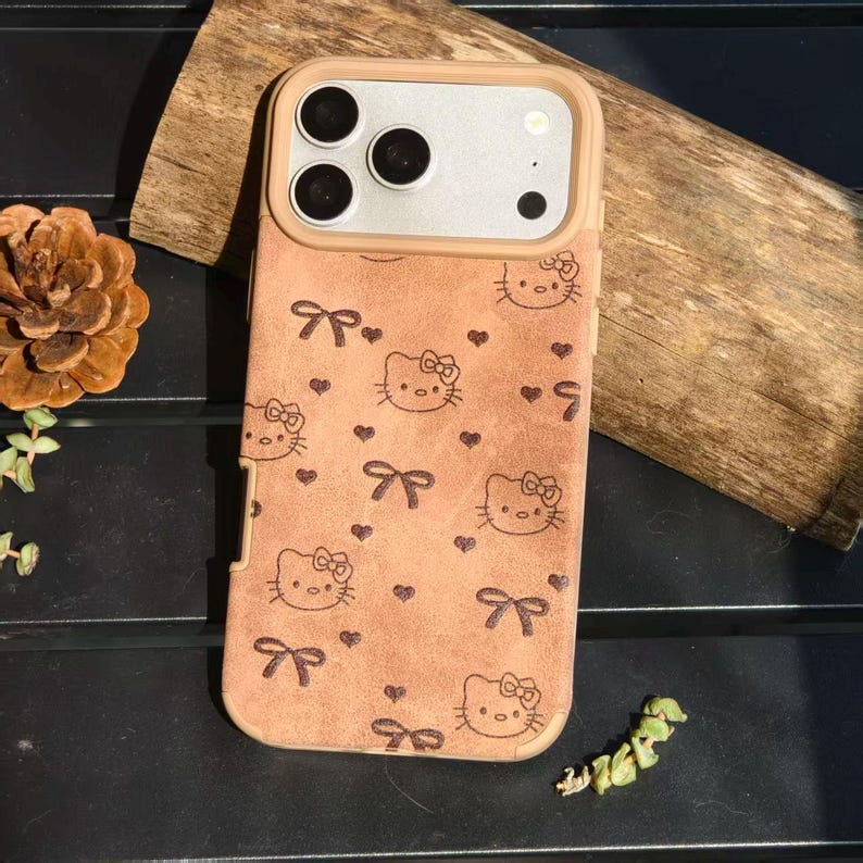 May include: A tan phone case featuring a Hello Kitty design with bows and hearts. The case has a tan frame around the camera and a light brown back. The Hello Kitty faces and bows are in a dark brown color.