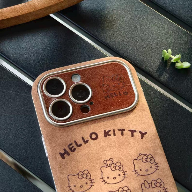 Op de afbeelding: Tan telefoonhoesje met Hello Kitty-ontwerpen. De hoes heeft een bruine cameralensrand en de woorden "HELLO KITTY" op de voorkant gedrukt. De hoes heeft een zachte, su&egrave;de-achtige textuur.