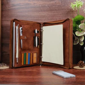 May include: A brown leather-look zipped portfolio organiser featuring a three-ring binder mechanism, a dedicated laptop sleeve, multiple pen loops, and several vertical slots for cards or documents. The interior includes a blank white paper pad.