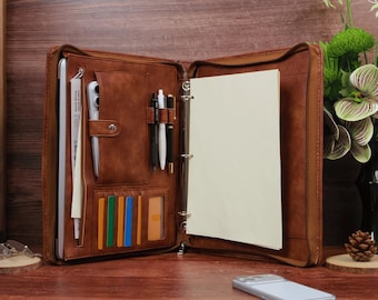 May include: A brown leather-like zippered portfolio organizer featuring a three-ring binder mechanism, a dedicated laptop sleeve, multiple pen loops, and several vertical slots for cards or documents. The interior includes a blank white paper pad.