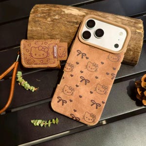 May include: A brown phone case and matching earbud case featuring a repeating pattern of Hello Kitty faces and bows. The phone case is designed for a smartphone with three camera lenses. The earbud case has a strap and the text "HELLO KITTY".