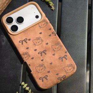 May include: A brown phone case featuring a repeating pattern of Hello Kitty faces, bows, and hearts. The case has a tan border around the camera and phone edges. The case is designed to fit a smartphone.