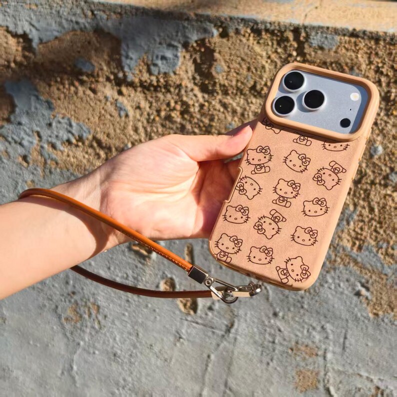 May include: A tan phone case featuring a repeating pattern of Hello Kitty characters in black outlines. The case includes a matching tan wrist strap attached with a silver metal clasp, designed to fit a smartphone with a triple-lens camera system.