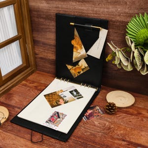Peut inclure: Un classeur noir à texture cuir avec un porte-stylo doré, doté d'une pochette triangulaire contenant une carte avec le texte A Special Letter, une photo de chat et une feuille à grille décorée de plusieurs petites photographies.