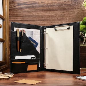 May include: A black portfolio folder featuring a three-ring binder mechanism, a metal clipboard clip, and multiple interior pockets. The pockets contain two pens, a passport, a white smartphone, and two small rectangular cards.