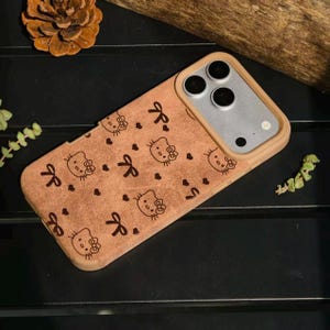 May include: A tan phone case featuring a repeating pattern of Hello Kitty faces, bows, and hearts. The case has a light brown border and a camera cutout. The phone case is designed to protect the phone.