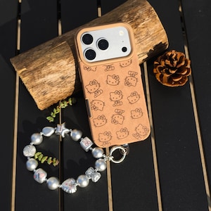 May include: A tan phone case featuring a repeating pattern of Hello Kitty characters. The case is paired with a silver and pearl bracelet with charms. A piece of wood and a pinecone are also in the image.