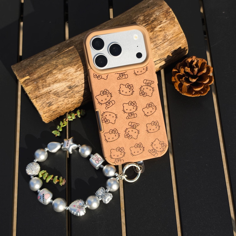 May include: A tan phone case featuring a repeating pattern of Hello Kitty characters. The case is paired with a silver and pearl bracelet with charms. A piece of wood and a pinecone are also in the image.