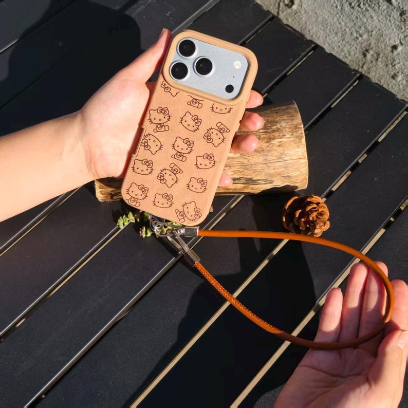 May include: A tan mobile phone case featuring a repeating pattern of black Hello Kitty character outlines. The case includes a raised camera cutout and a long, thin brown wrist strap attached to the bottom with a silver metal clasp.