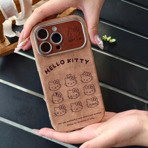 Op de afbeelding: Bruine telefoonhoes met Hello Kitty-ontwerpen. De hoes heeft een bruine leerachtige textuur met de woorden "HELLO KITTY" en meerdere Hello Kitty-illustraties. De camera heeft een bruin frame met het woord "HELLO".