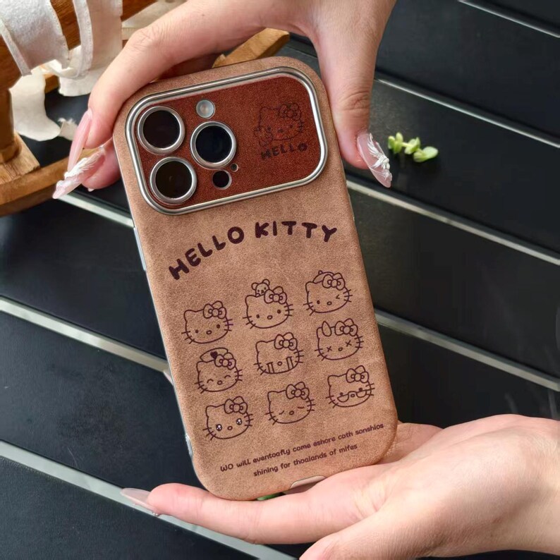Op de afbeelding: Bruine telefoonhoes met Hello Kitty-ontwerpen. De hoes heeft een bruine leerachtige textuur met de woorden "HELLO KITTY" en meerdere Hello Kitty-illustraties. De camera heeft een bruin frame met het woord "HELLO".