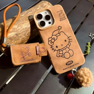 May include: Tan phone case and wallet set featuring a Hello Kitty design. The phone case has a winter-themed Hello Kitty graphic and the text "Good Winter" and "Have a good time". The wallet has a Hello Kitty graphic on a skateboard.