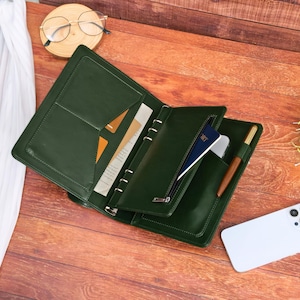 May include: A dark green leather-like planner organizer featuring a six-ring metal binder mechanism, multiple interior pockets, a zippered compartment containing a blue passport, and a pen loop holding a brown pen.