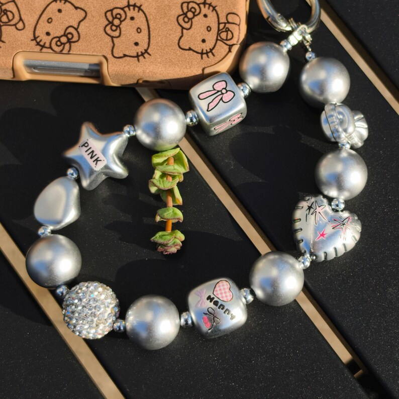 May include: Silver beaded bracelet with charms, including a star with "PINK" text, a heart, and a cube with a bow design. The bracelet also features a cluster of clear rhinestones and a cube with the word "Happy" and a heart design.