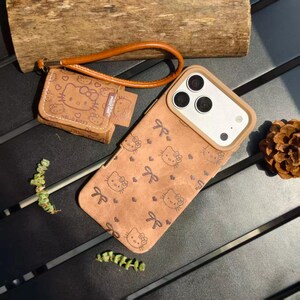 May include: Brown phone case and matching small wallet featuring a repeating pattern of Hello Kitty faces and bows. The phone case has a tan border around the camera. The wallet has a wrist strap. The text "HELLO KITTY" is visible on the wallet.