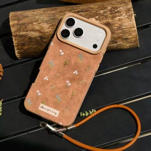 Puede incluir: Una funda de teléfono de color marrón claro con un diseño floral y una correa de cuero marrón. La funda tiene forma rectangular con un recorte para la cámara y una pequeña etiqueta que dice "Beautiful Story". La correa está sujeta a la parte inferior de la funda con un cierre plateado.
