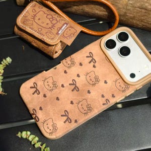 May include: Tan phone case and matching wallet featuring a repeating pattern of Hello Kitty faces, bows, and hearts. The phone case has a tan border and camera cutouts. The wallet has a wrist strap and the words "Hello Kitty" printed on it.