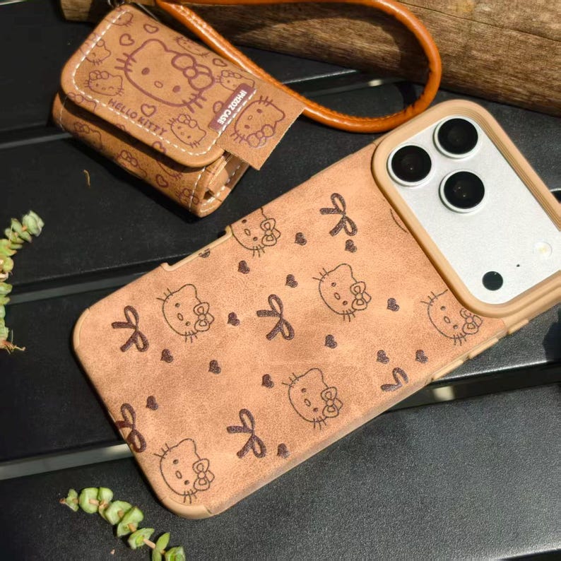 May include: Tan phone case and matching wallet featuring a repeating pattern of Hello Kitty faces, bows, and hearts. The phone case has a tan border and camera cutouts. The wallet has a wrist strap and the words "Hello Kitty" printed on it.