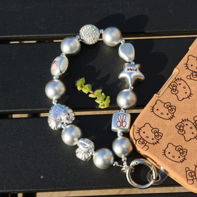 May include: A silver beaded wristlet with various charms, including a star with "PINK" text, a heart, and a bunny. The wristlet is next to a brown phone case with a repeating Hello Kitty pattern.