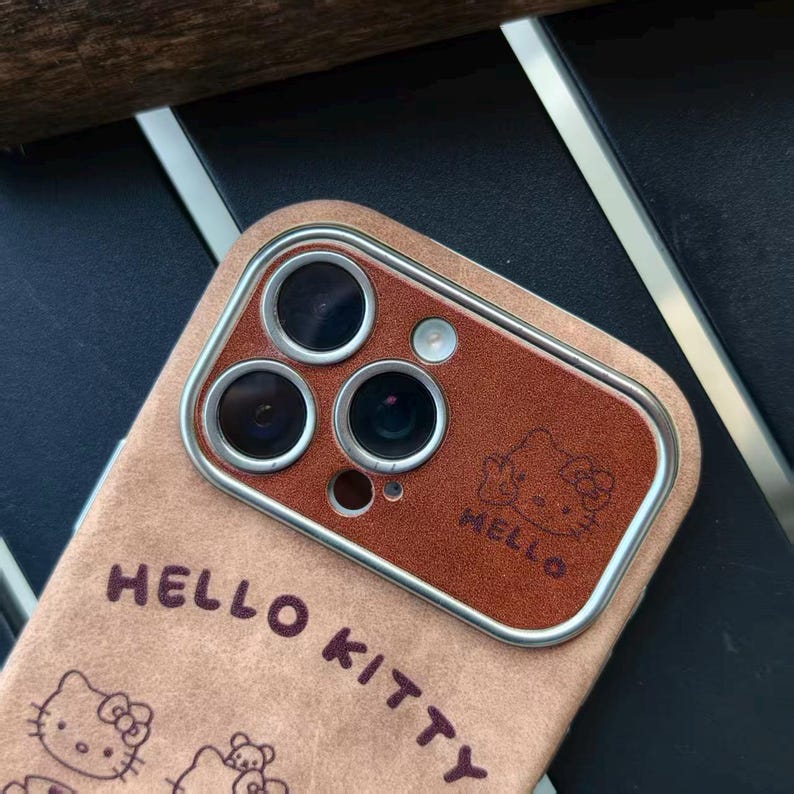 Op de afbeelding: Een tan telefoonhoesje met een bruine cameralens cover. De hoes heeft de woorden "HELLO KITTY" en een Hello Kitty-afbeelding. De cameralens cover heeft het woord "HELLO" en een Hello Kitty-afbeelding.